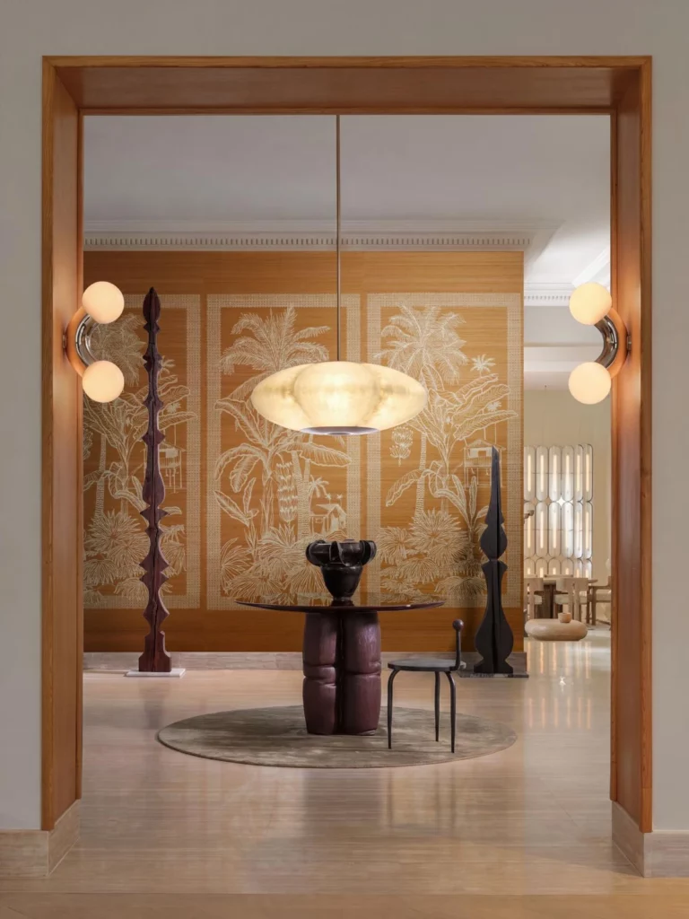 Arte Nilaya: goldfarbenes Palmenpanorama mit stilisierten Hütten hinter einem runden Tisch, durch eine offene Doppeltür betrachtet.