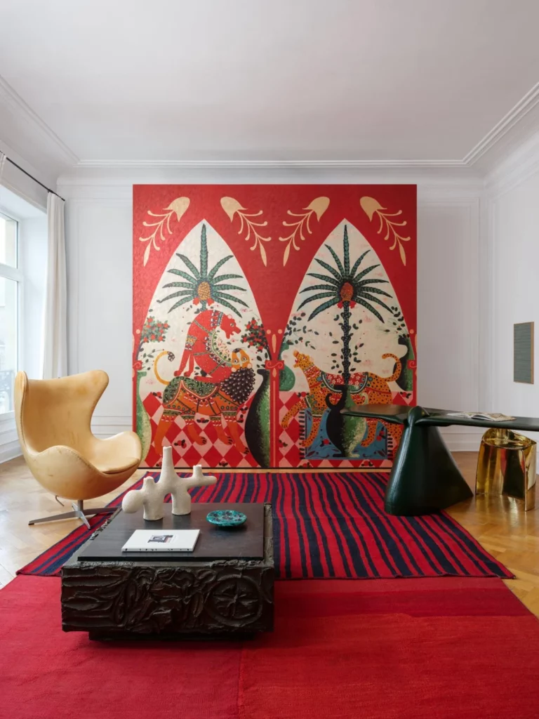 Arte Armalu: rotes, gerahmtes Wandbild mit Figuren und Pflanzen hinter einer niedrigen Sitzbank.