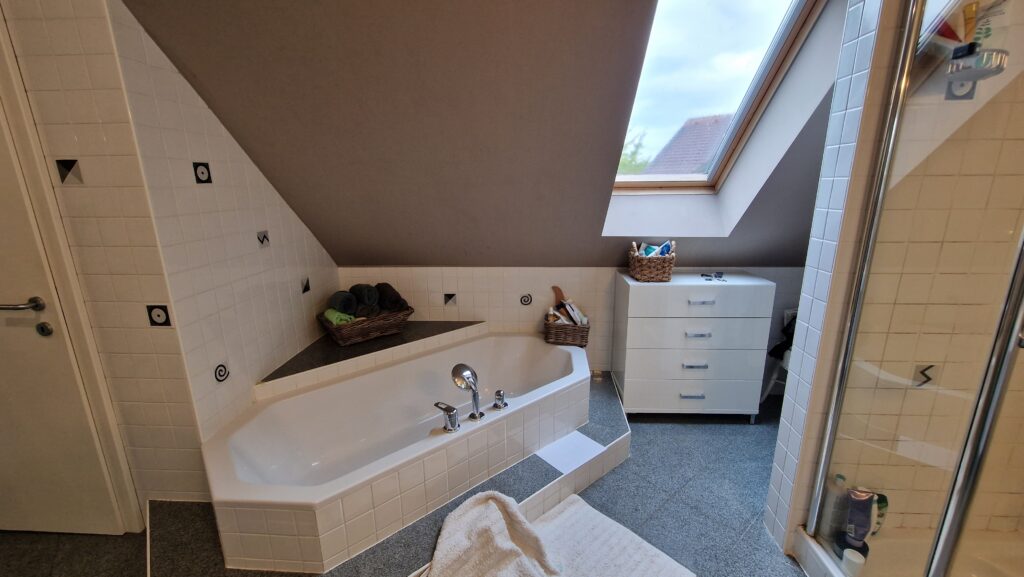 Badezimmer vor dem Umbau mit eingebauter Badewanne, gefliesten Wänden und einem Dachfenster.