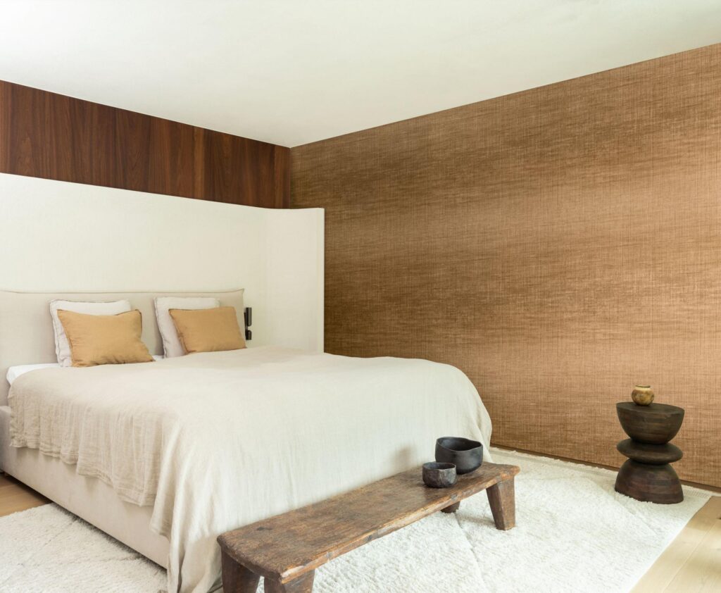Arte Fade: braune Strukturwand hinter einem hell bezogenen Bett und einer niedrigen Holzbank.