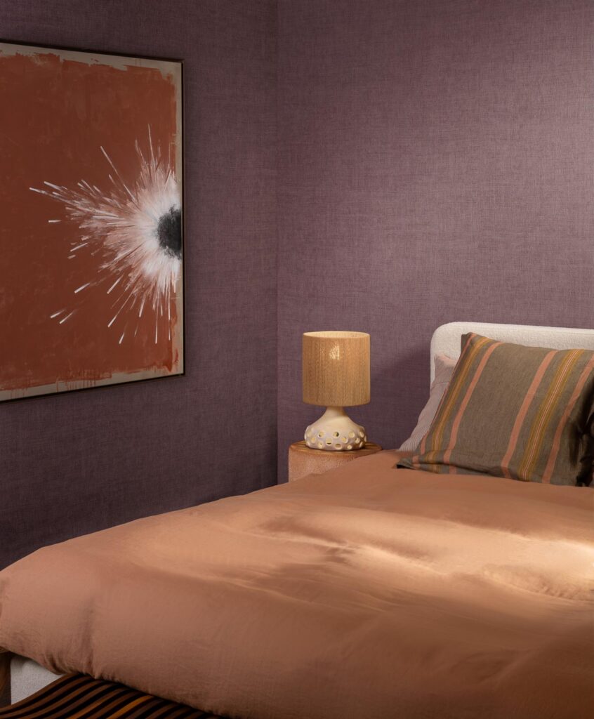 Arte Mandu: violettbraune Strukturwand hinter einem Bett mit warmrosa Bettwäsche.