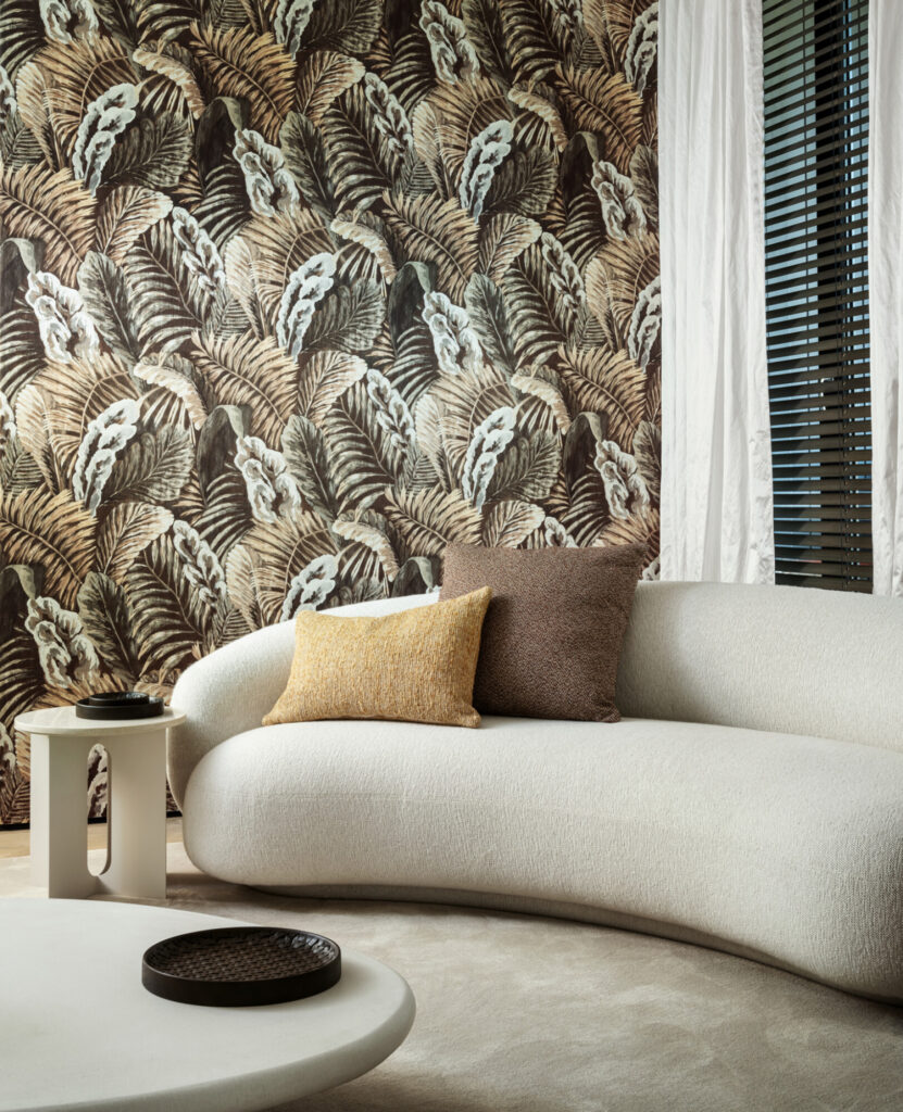 Arte Verdure: dichtes Palmenmotiv in Natur- und Brauntönen hinter einem geschwungenen hellen Sofa.