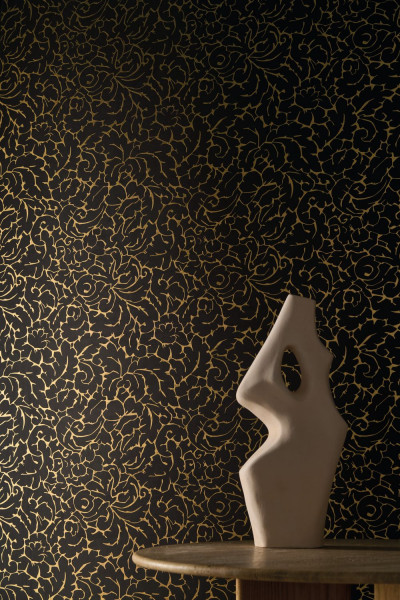 Dunkle Wand mit feiner goldfarbener Pflanzenzeichnung hinter einer hellen Skulptur.