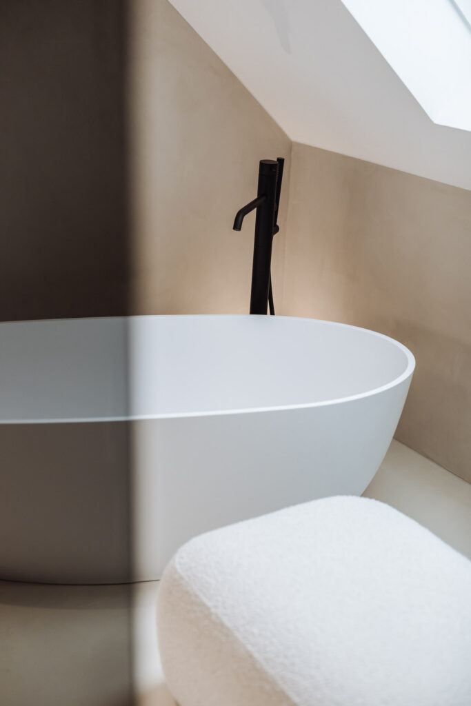 Detail der weißen Badewanne vor einer beigefarbenen Wand mit dunkler Armatur und hellem Hocker.
