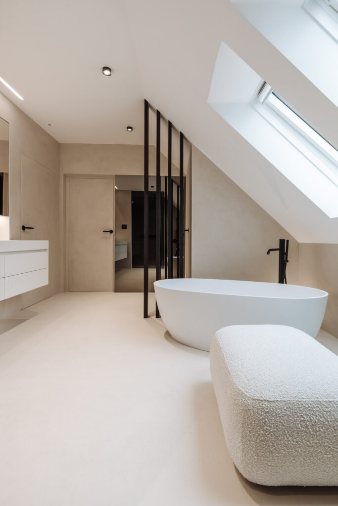 Freistehende weiße Badewanne unter dem Dachfenster, daneben dunkle Lamellen und ein heller Hocker.