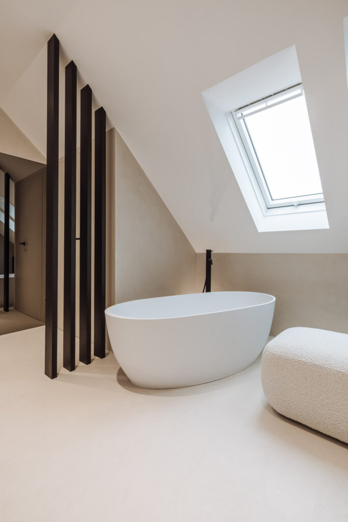 Weiße ovale Badewanne unter einem Dachfenster neben einem dunklen Lamellenteiler.