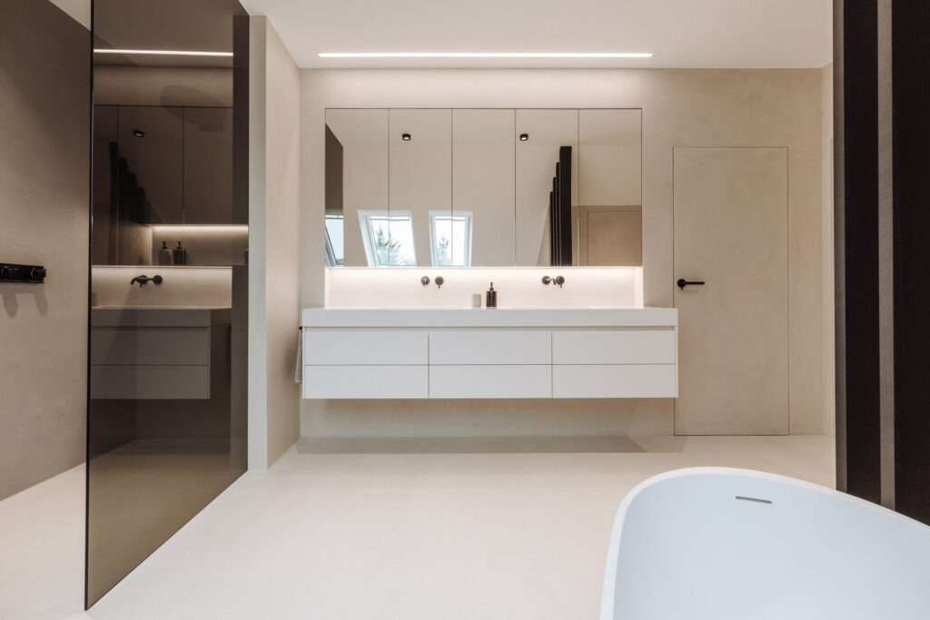 Blick von der Badewanne auf den weißen Waschtisch, den großen Spiegel und die seitliche Glasabtrennung.