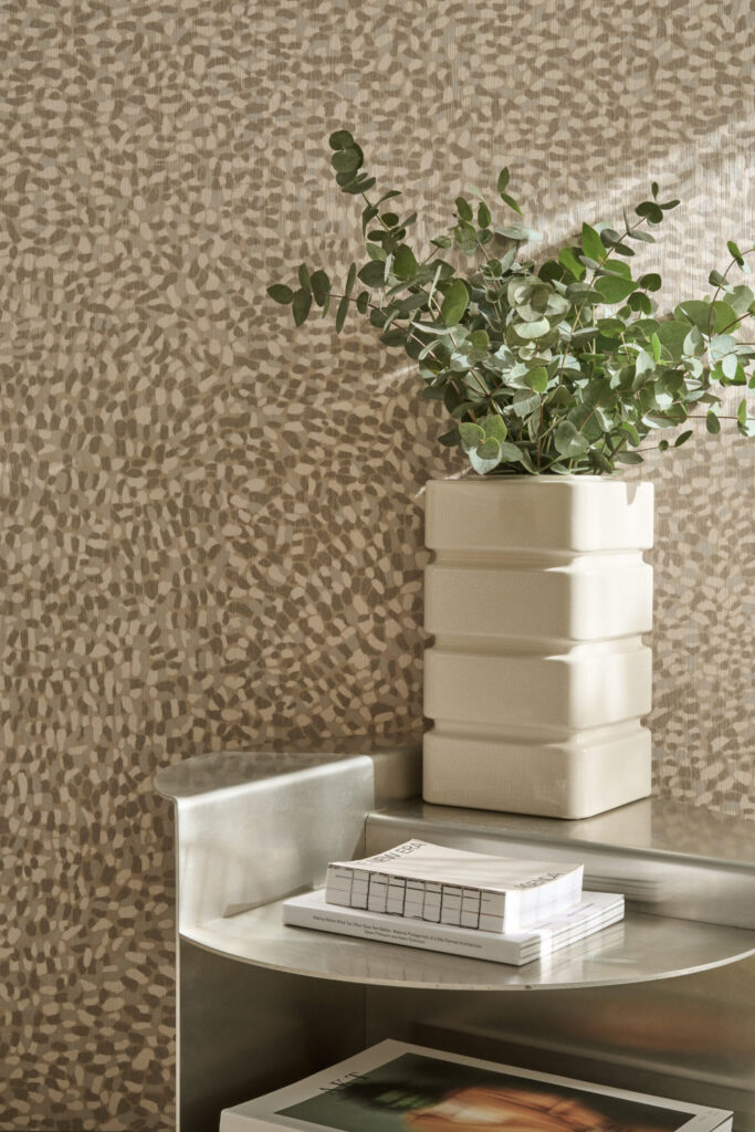 Kleinteilig gesprenkeltes Wandmuster in Beige und Braun hinter einer weißen Vase mit grünen Zweigen.