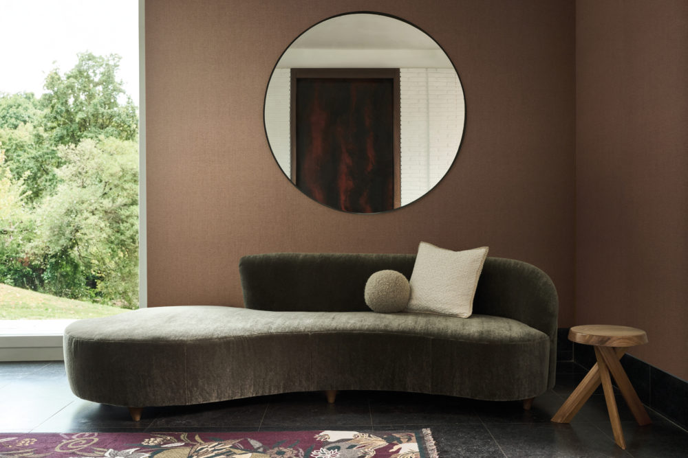Ruhige braune Wand mit rundem Spiegel hinter einem geschwungenen dunkelgrünen Sofa.