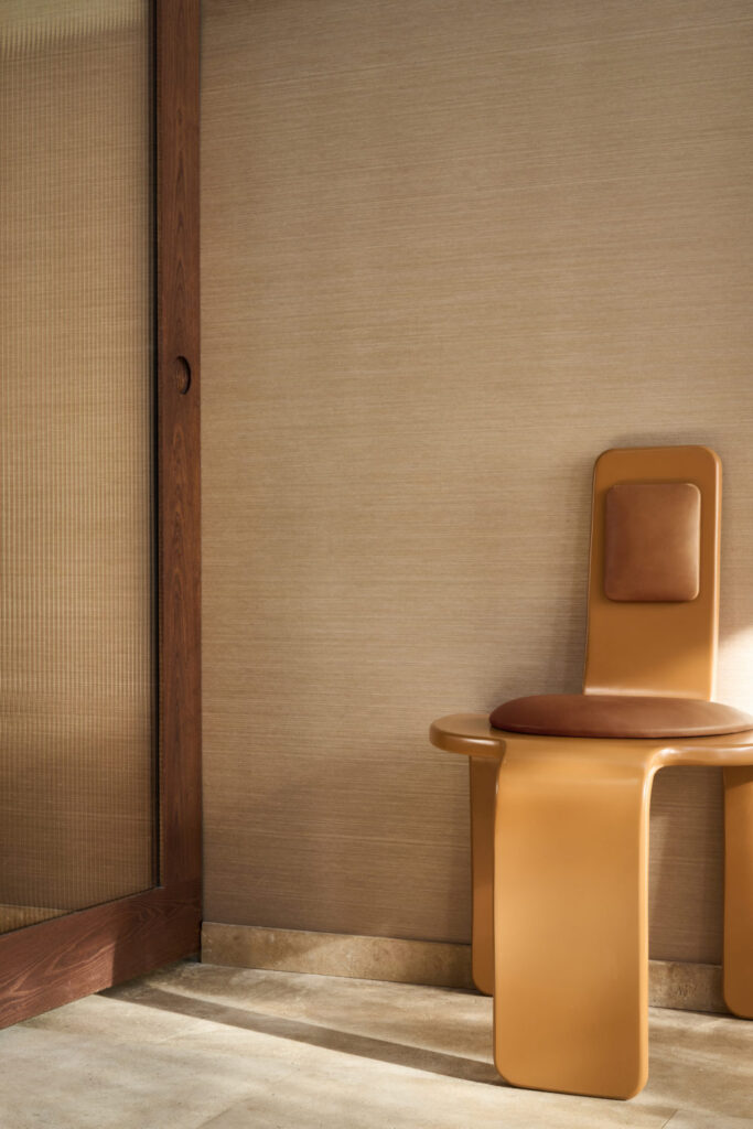 Fein strukturierte beige Wandbekleidung neben einer Holztür und einem skulpturalen ockerfarbenen Stuhl.