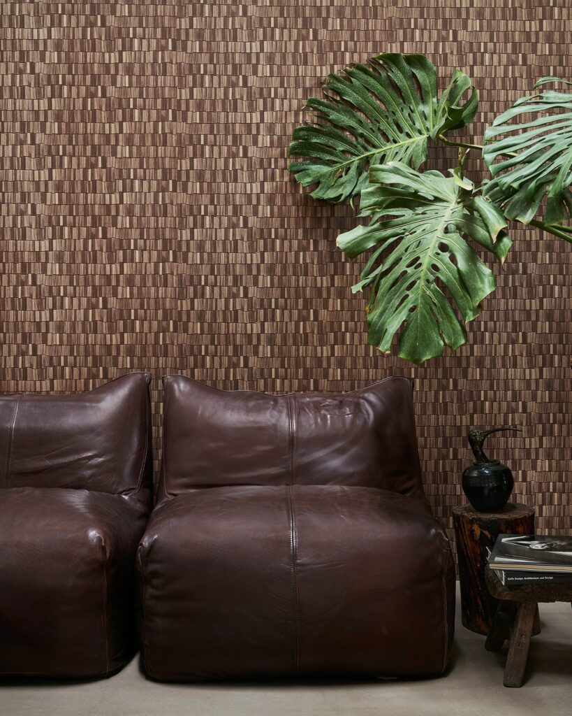 Braune strukturierte Wandbekleidung Tavillon hinter einem Ledersofa mit grünen Blättern – Wall&decò Essential Wallpaper 2025