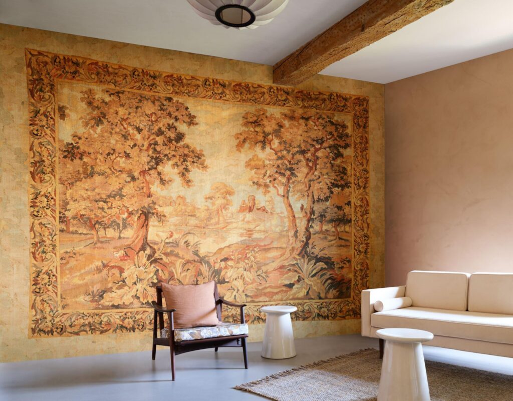 Arte Escapade: warmtoniges Landschaftsdekor mit ornamentaler Einfassung hinter einem Sessel und einem hellen Sofa.