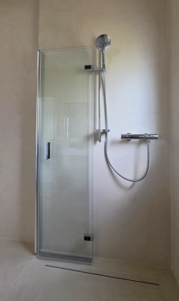 An die Wand gefaltete Duschglaselemente neben Handbrause und Armatur; darunter eine schmale Ablaufrinne.