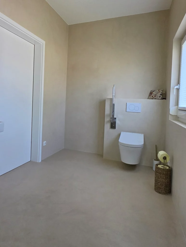 Wandhängendes WC mit hochgeklapptem Stützgriff neben dem Fenster und einer weißen Tür.