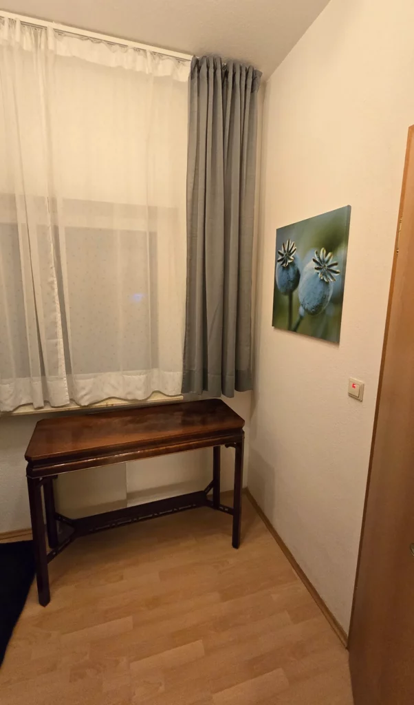 Fenster mit Vorhängen und kleiner Holzkonsole im früheren Vorraum.