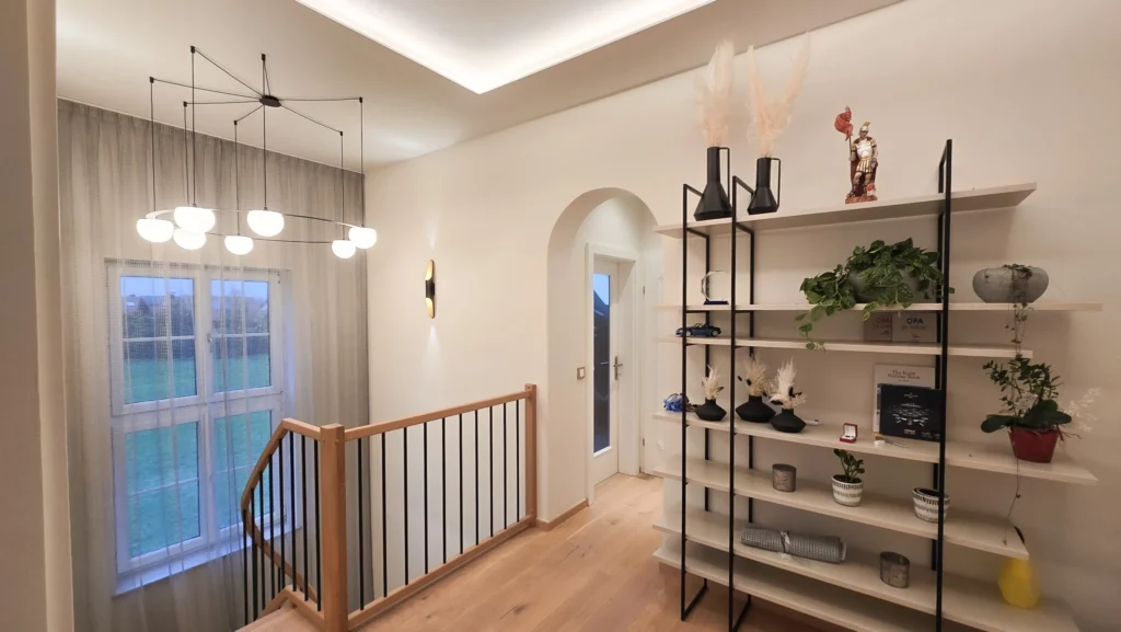 Helle Galerie mit hohem Fenster, mehrteiliger Pendelleuchte, Holzgeländer und offenem Regal.