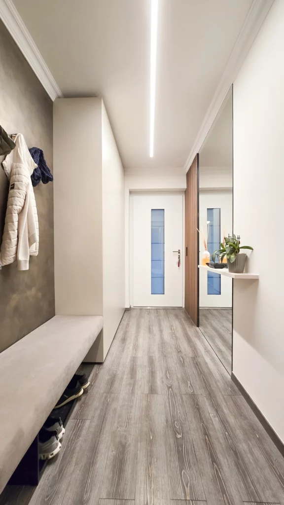 Helle Garderobe mit Sitzbank, großer Spiegelfläche, Holzlamellen und langer Lichtlinie an der Decke.