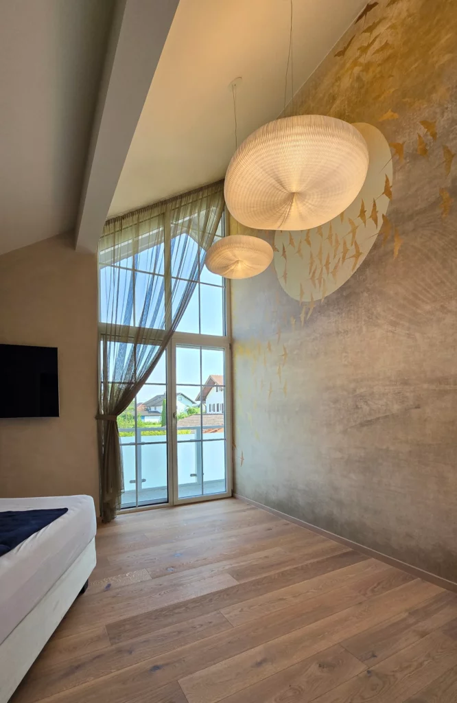 Schlafzimmer mit goldfarbenem Vogelmotiv an der Wand, zwei weißen Pendelleuchten und hohem Fenster mit Vorhang.