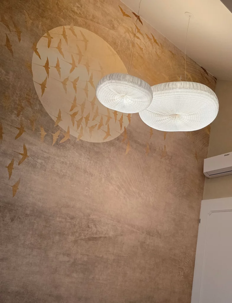 Goldfarbenes Vogelmotiv mit hellem Kreis an einer strukturierten Schlafzimmerwand hinter zwei weißen Pendelleuchten.