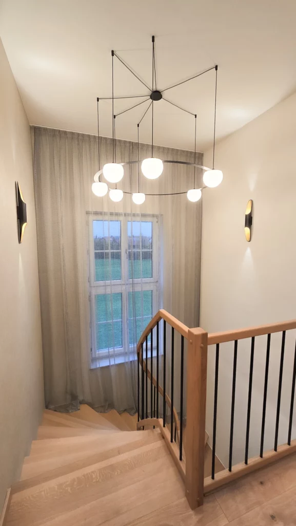 Treppenabgang mit Holzgeländer vor einem hohen Fenster und einer Pendelleuchte mit mehreren weißen Schirmen.