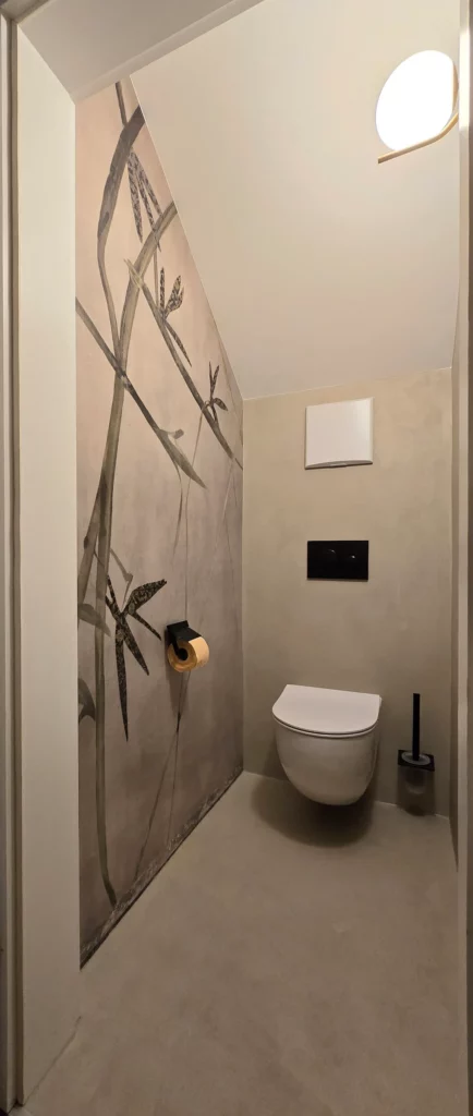 Schmales WC unter einer Schräge mit großem Bambusmotiv an der linken Wand und warmer Wandleuchte.
