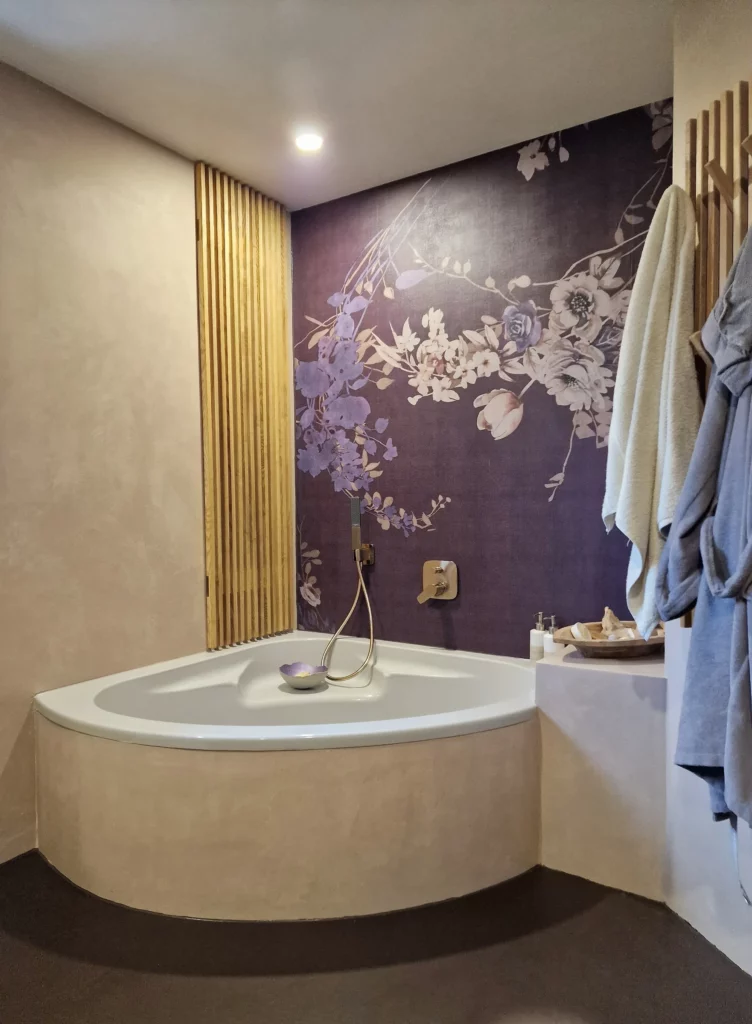 Eckbadewanne vor einer violetten Wand mit Blütenmotiv, daneben senkrechte Holzleisten und beige Wandflächen.