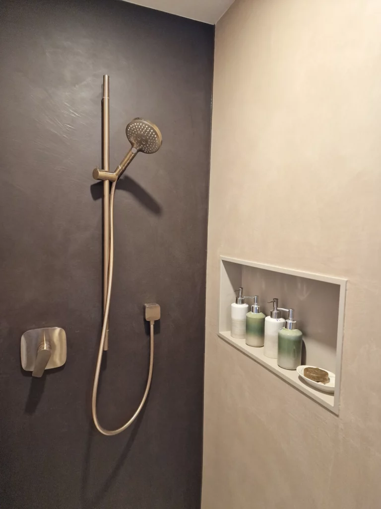 Duscharmatur an einer dunklen Wand neben einer hellen Ablagenische mit Seifenspendern.