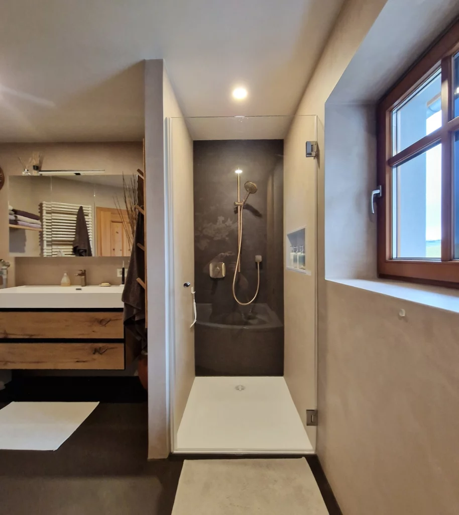 Dusche mit Glasabtrennung, dunkler Rückwand und weißer Duschfläche zwischen Waschtisch und Fenster.