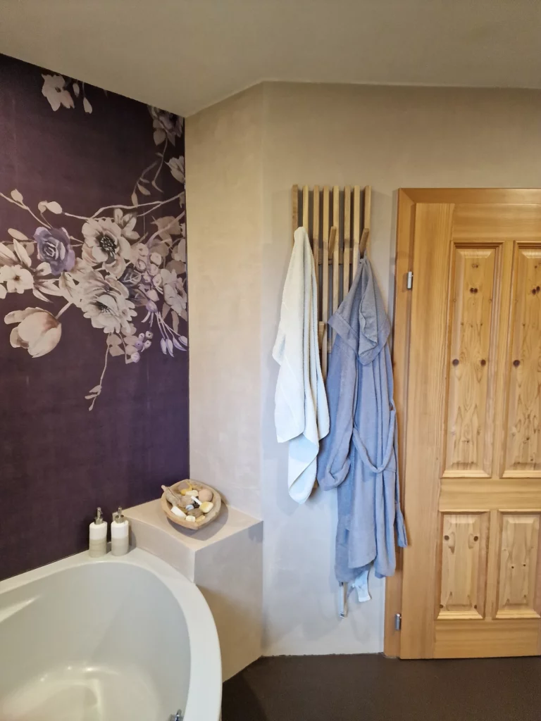 Violette florale Motivwand neben einer beigen Wand mit senkrechter Hakenleiste, Handtüchern und einer Holztür am Wannenbereich.