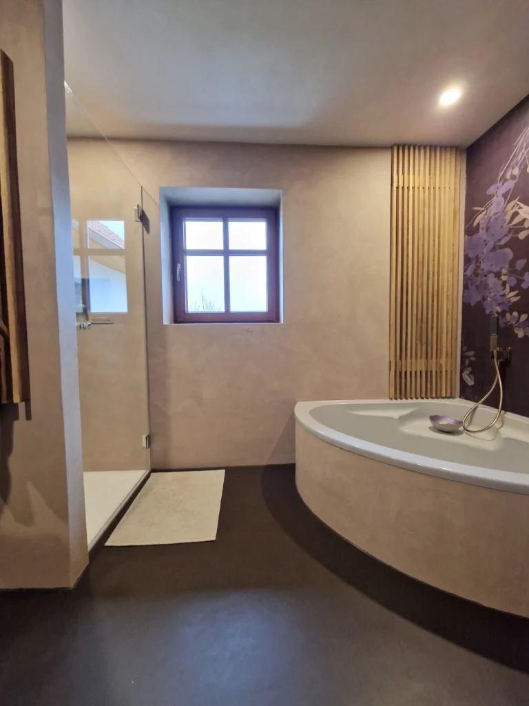 Fenster zwischen der verglasten Dusche und der Eckbadewanne, umgeben von beigen Wandflächen und einer violetten floralen Motivwand.