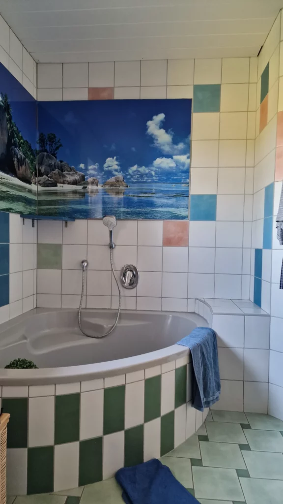 Eckbadewanne vor weißen und farbigen Fliesen mit einem Strandmotiv an der Wand, vor der Neugestaltung.