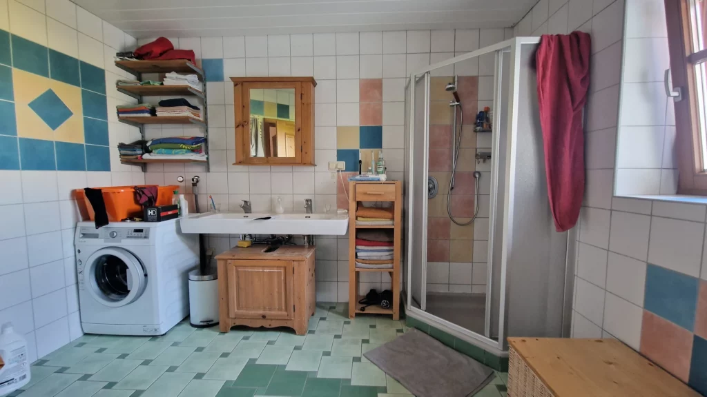 Waschbereich mit Spiegel, offenen Regalen und Waschmaschine neben einer Duschkabine im gefliesten Badezimmer vor der Neugestaltung.
