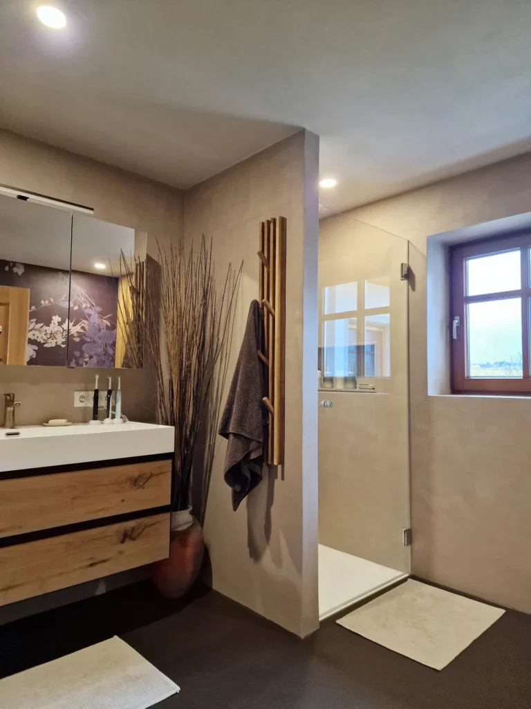 Waschtisch mit Holzfronten neben einer beigen Trennwand mit Hakenleisten; rechts die verglaste Dusche.
